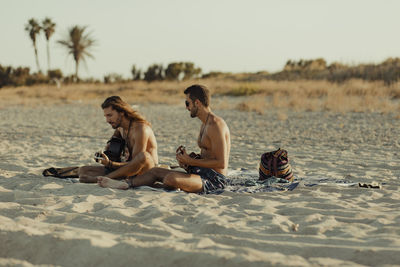 Relaxed talented man playing acoustic guitar for male friend while sitting together on sandy beach at sunset