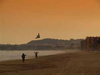 Friends flying kite at beach against sky during sunset