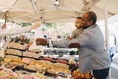 Man with daughter buying fruit and vegetables from female seller in market stall