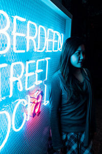 Young woman standing by illuminated blue sign in dark at night