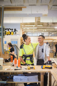 Cheerful female trainee standing with arm around male coworker at workbench