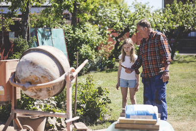 Grandfather giving shovel to granddaughter while standing by cement mixer in yard