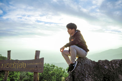 Side view of man sitting on rock against sky