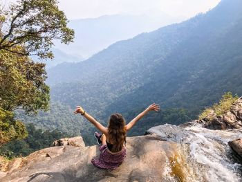 Rear view of woman with arms raised sitting on cliff