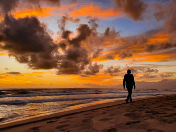 Rear view of man walking at beach against sky during sunset