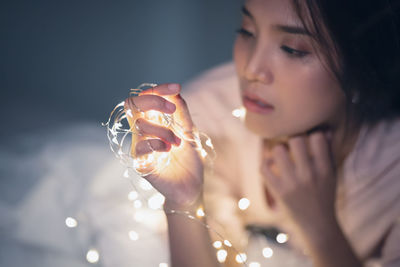 Young woman holding illuminated string light on bed at home