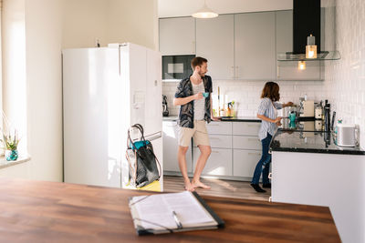 Man drinking coffee while looking at woman working in kitchen