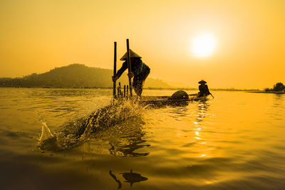 Fishermen on rowboat in sea against orange sky during sunset