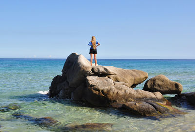 Woman standing on rock by sea against clear blue sky