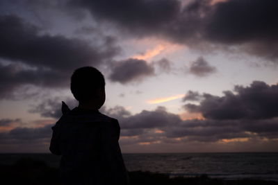 Silhouette boy standing at beach against cloudy sky during sunset