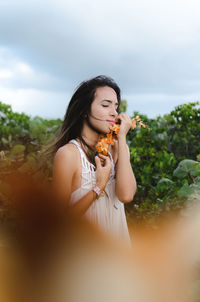 Young woman holding orange flowers against trees against sky