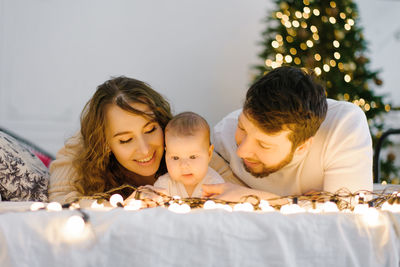 Happy mom and dad lie on the bed next to their baby in the background of christmas lights