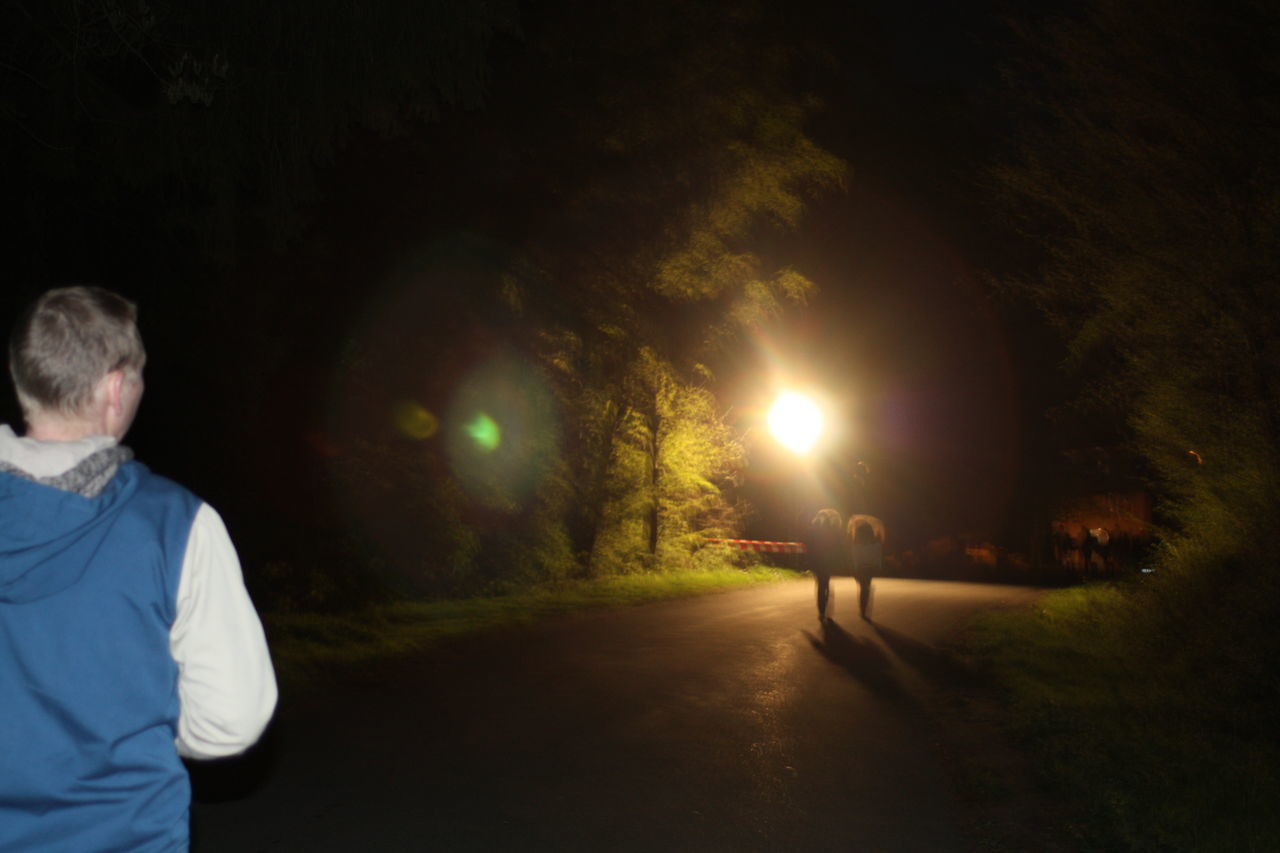 REAR VIEW OF MAN WALKING ON ILLUMINATED ROAD AT NIGHT