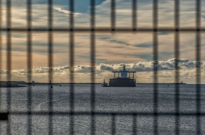 Boat moving in sea seen through fence against cloudy sky during sunset