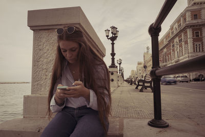 Young woman using phone while sitting in city against sky