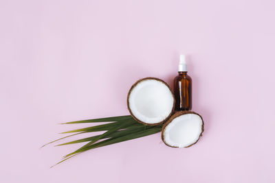 Flat lay with cut halves of coconut and green palm leaf, essential oil bottle on pink background