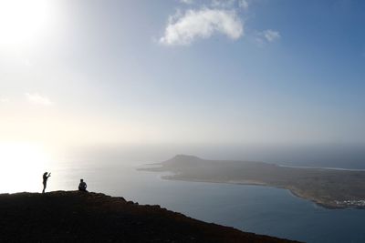Silhouette friends on mountain with sea in background against blue sky