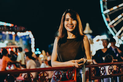 Portrait of smiling young woman standing by railing in city at night