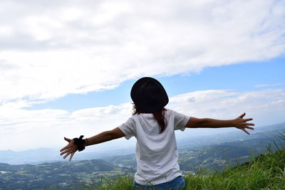 Rear view of woman with arms outstretched on grassy hill against cloudy sky