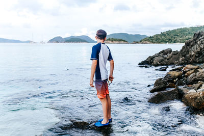 Rear view of man standing on rock at sea shore against sky