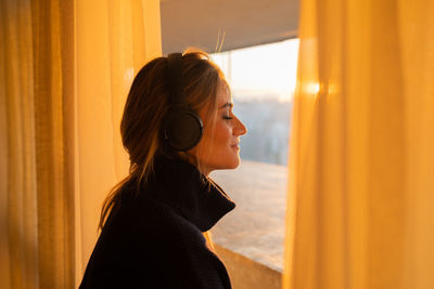 Side view of young woman listening to music with headphones by the window. city view landscape