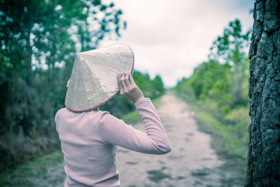 Rear view of woman wearing asian style conical hat standing by tree trunk on field