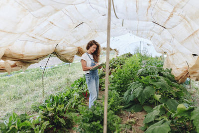 Smiling farm worker with clipboard taking vegetables inventory in greenhouse