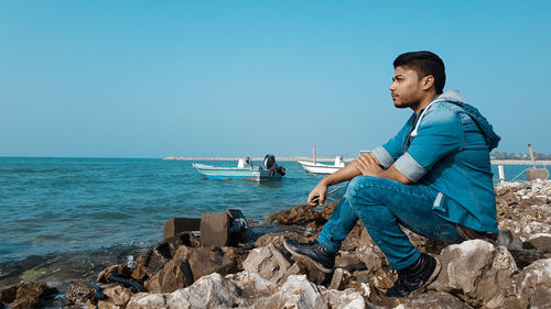 Side view of thoughtful man sitting on rocks by sea against clear sky
