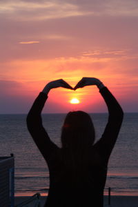 Rear view of woman making heart shape against orange sky over sea during sunset