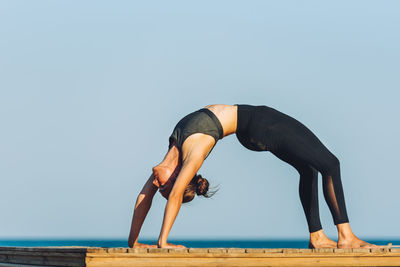 Side view of woman practicing wheel pose on boardwalk against sea