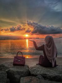 Rear view of woman sitting at beach against sky during sunset