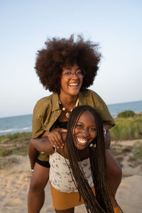 Cheerful young african american female with afro pigtails giving piggyback ride to laughing girlfriend with curly hair while having fun during summer holidays together on seashore