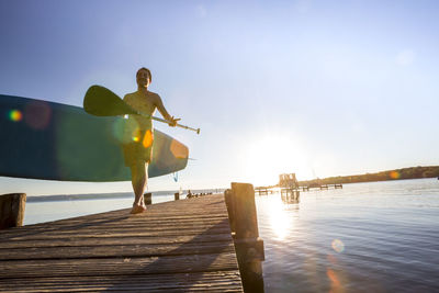 Shirtless man walking with paddleboard on pier over lake against sky