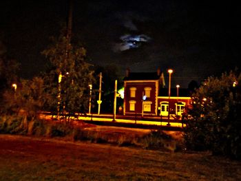 Illuminated street by building against sky at night