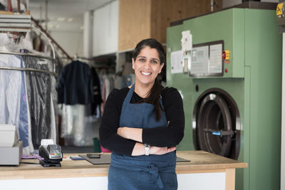 Portrait of smiling confident mature female dry cleaner standing with arms crossed at laundromat