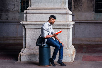 Side view of young black man sitting on street in new york city, reading red book.
