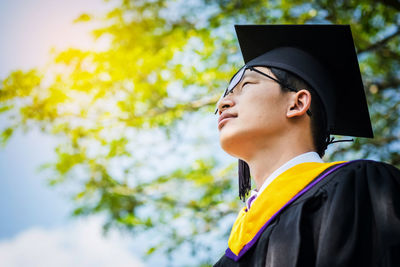 Low angle view of young man in graduation gown standing against sky