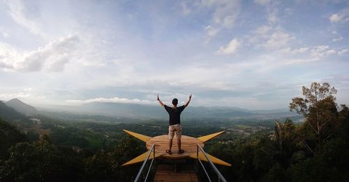 Rear view of man with arms raised standing at observation point against sky