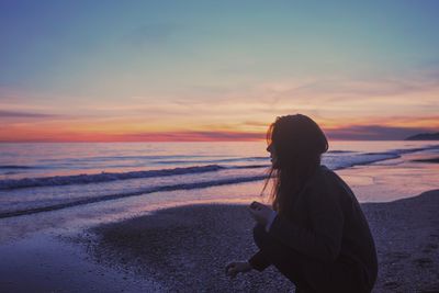 Woman crouching at beach against sky during sunset