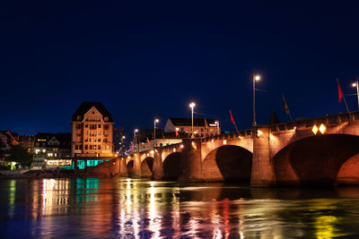 Illuminated bridge over river by buildings against sky at night