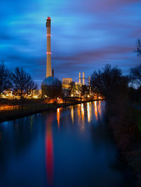 Illuminated factory by river against sky