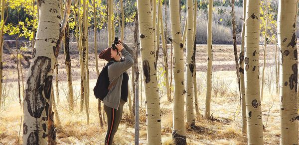 Side view of woman photographing while standing by tree trunk