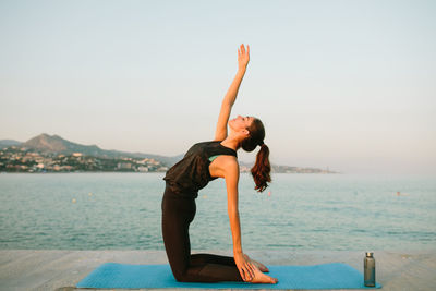 Side view of tranquil female in ustrasana practicing yoga with closed eyes and raised arm on pier near sea during sundown in summer