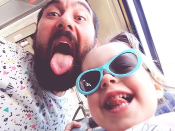 Low angle portrait of playful father and daughter sticking out tongues by window