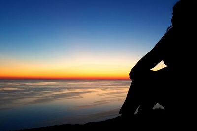 Silhouette woman sitting at seashore against clear sky during sunset