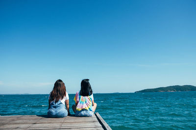 Rear view of friends sitting on pier over sea against clear blue sky