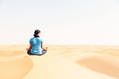 Rear view of man sitting on sand at beach against clear sky