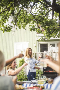 Mature woman preparing food on barbecue while toasting wineglasses with friends at backyard party