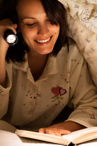 Smiling woman holding flashlight reading book on bed under blanket