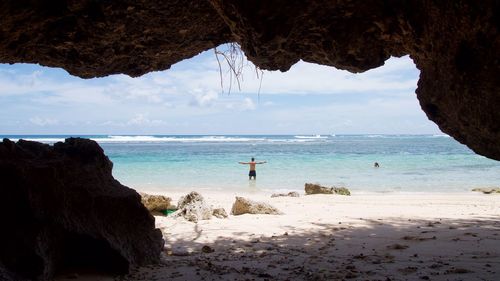 Man with arms outstretched at beach against sky seen through cave